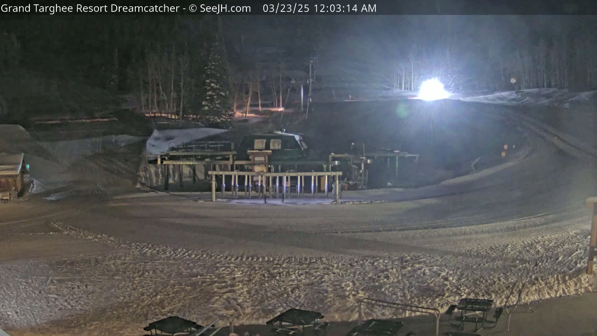 A nighttime view of a snow-covered ski slope with a lift structure and some outdoor furniture in the foreground under artificial light.