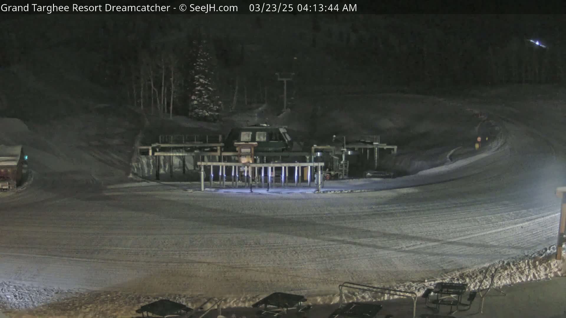 A snow-covered ski slope at night, illuminated by artificial lights, shows a ski lift and a small building.