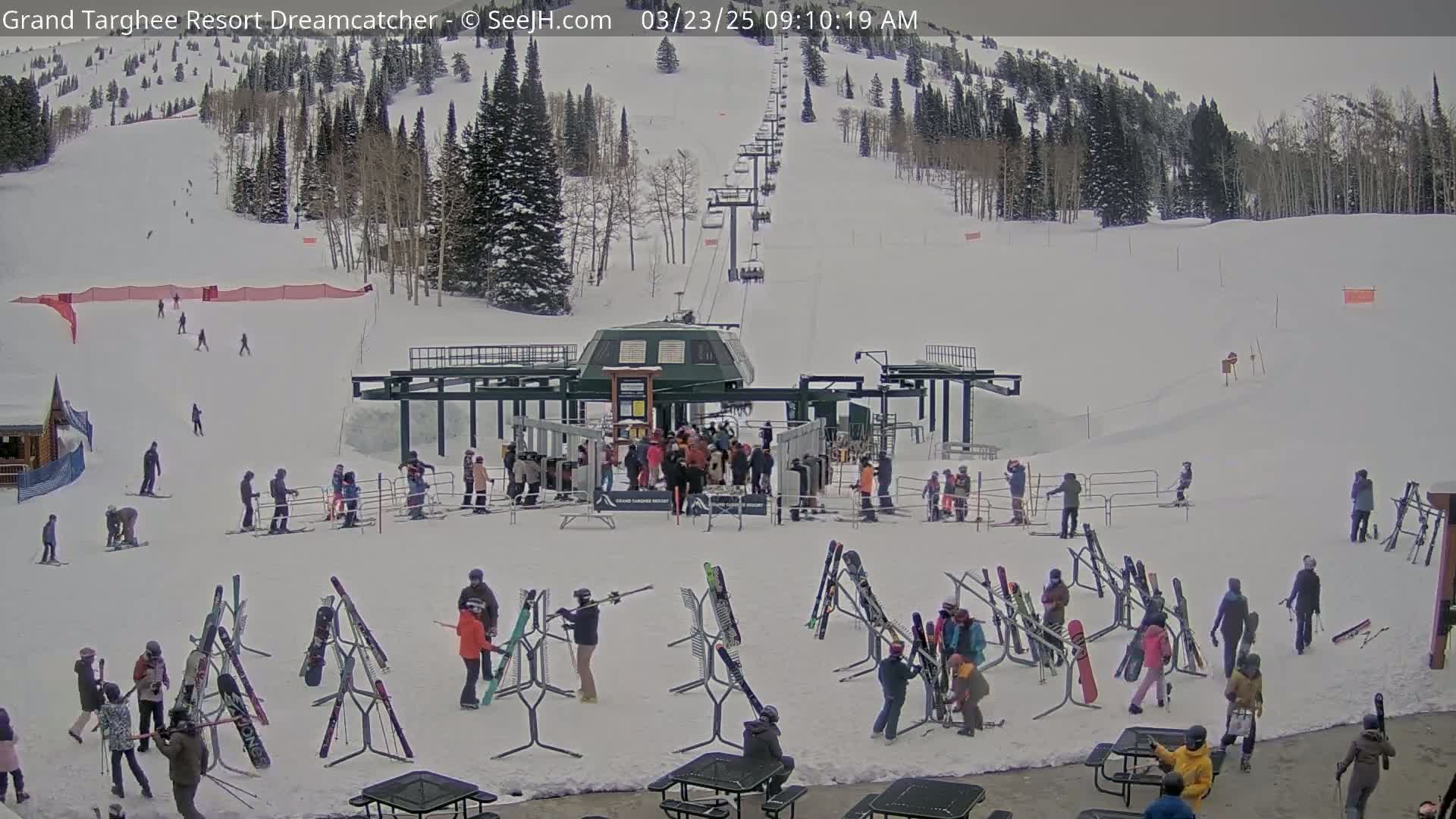 A ski lift base area with numerous skiers and snowboarders, many skis and snowboards on racks, and a snow-covered mountainside in the background under an overcast sky.