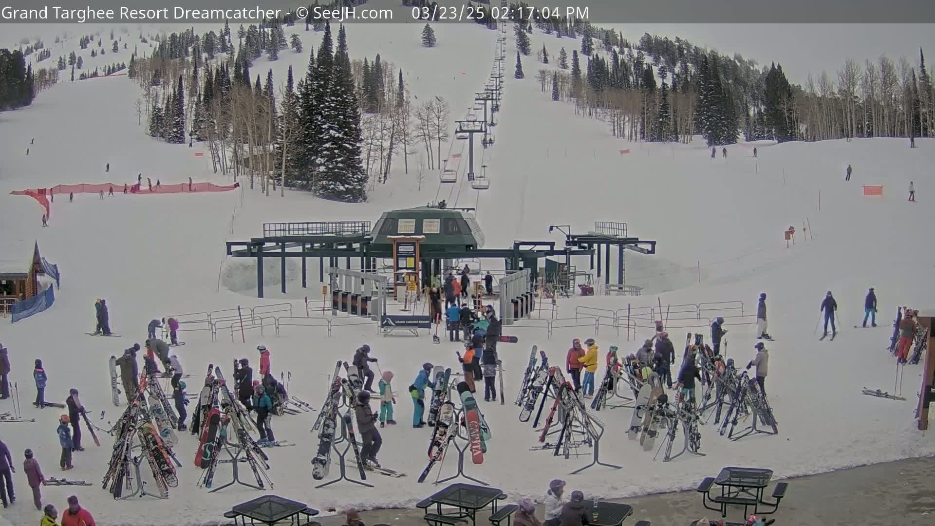 A ski lift base area with numerous skiers and snowboarders around racks of equipment, under an overcast sky on a snow-covered mountain.