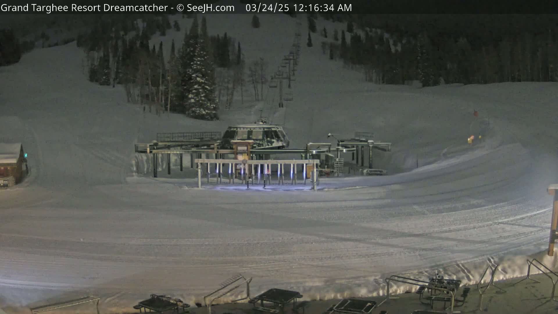 A snow-covered ski slope at night, illuminated by artificial lights, features a chairlift and its base station.