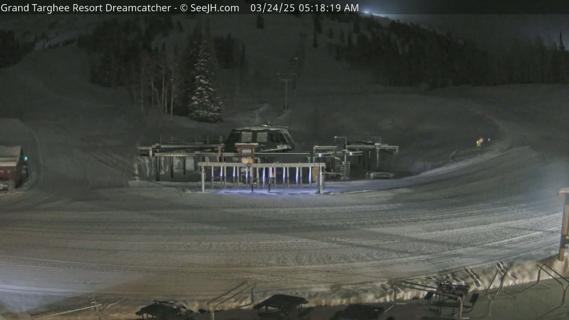 A snow-covered ski slope at night, illuminated by artificial lights, features a ski lift and a snowcat near the base of the hill.