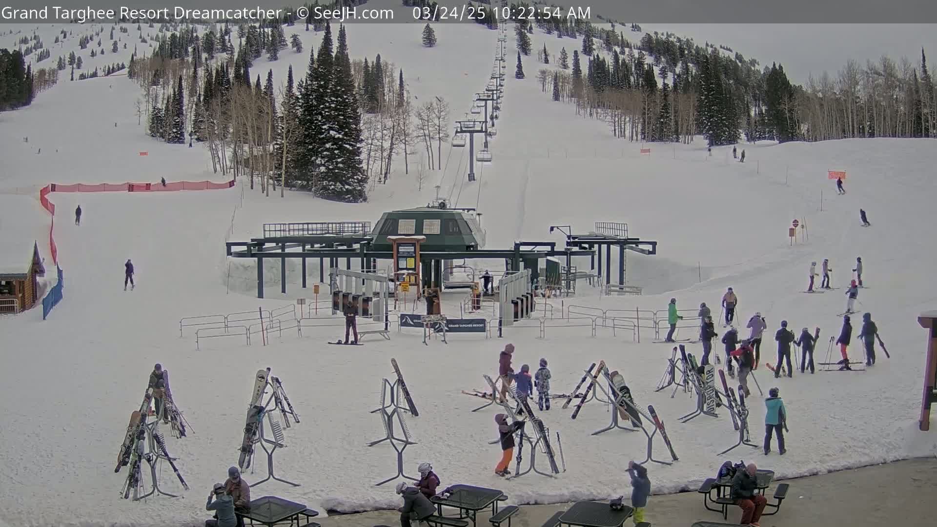 A ski resort base area with a chairlift, skiers and snowboarders, and several ski racks on a snowy day.