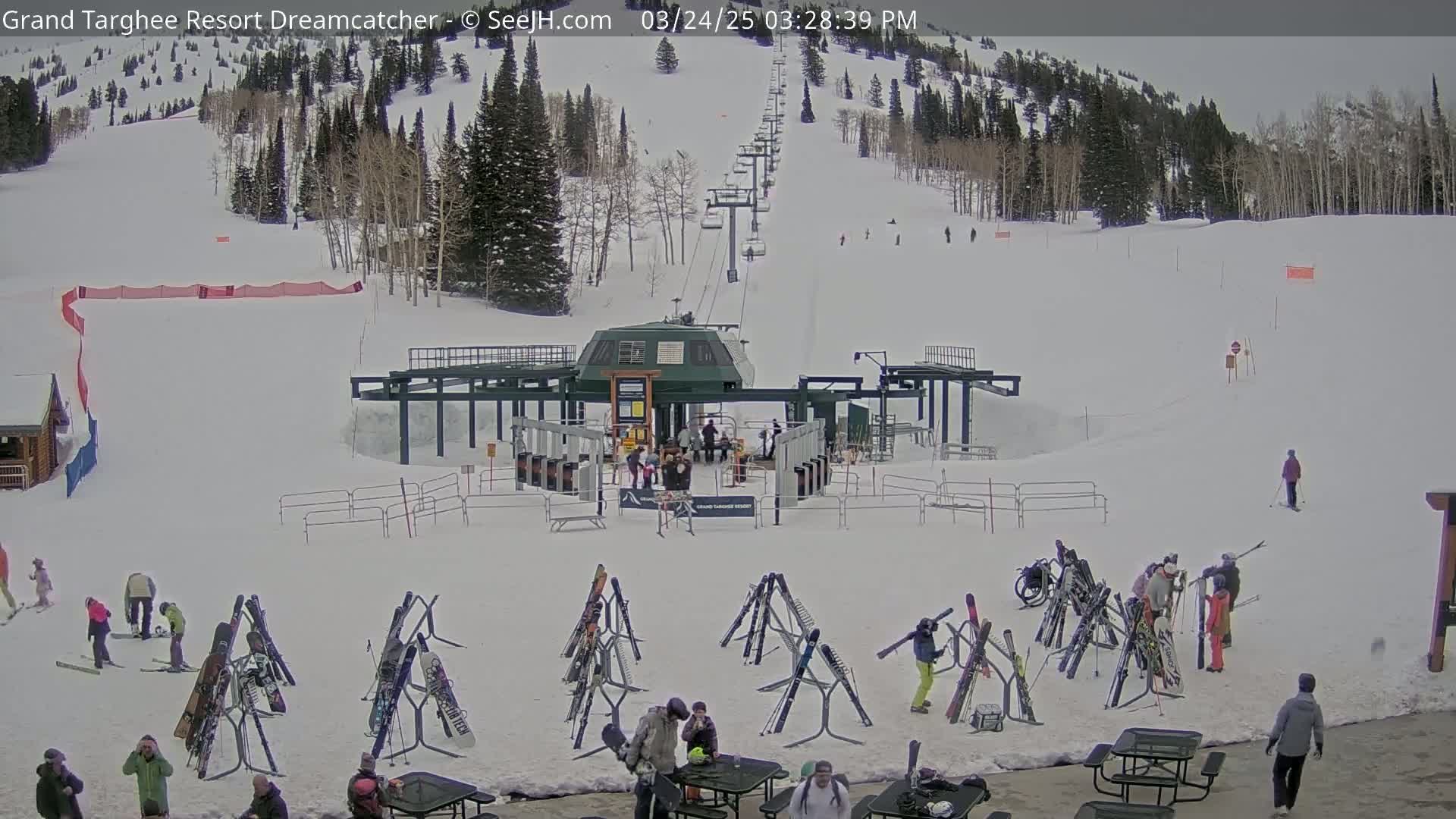 A ski lift base area with skiers and snowboarders, many skis and snowboards on racks, and several people sitting at picnic tables in the snow, on a partly cloudy day.