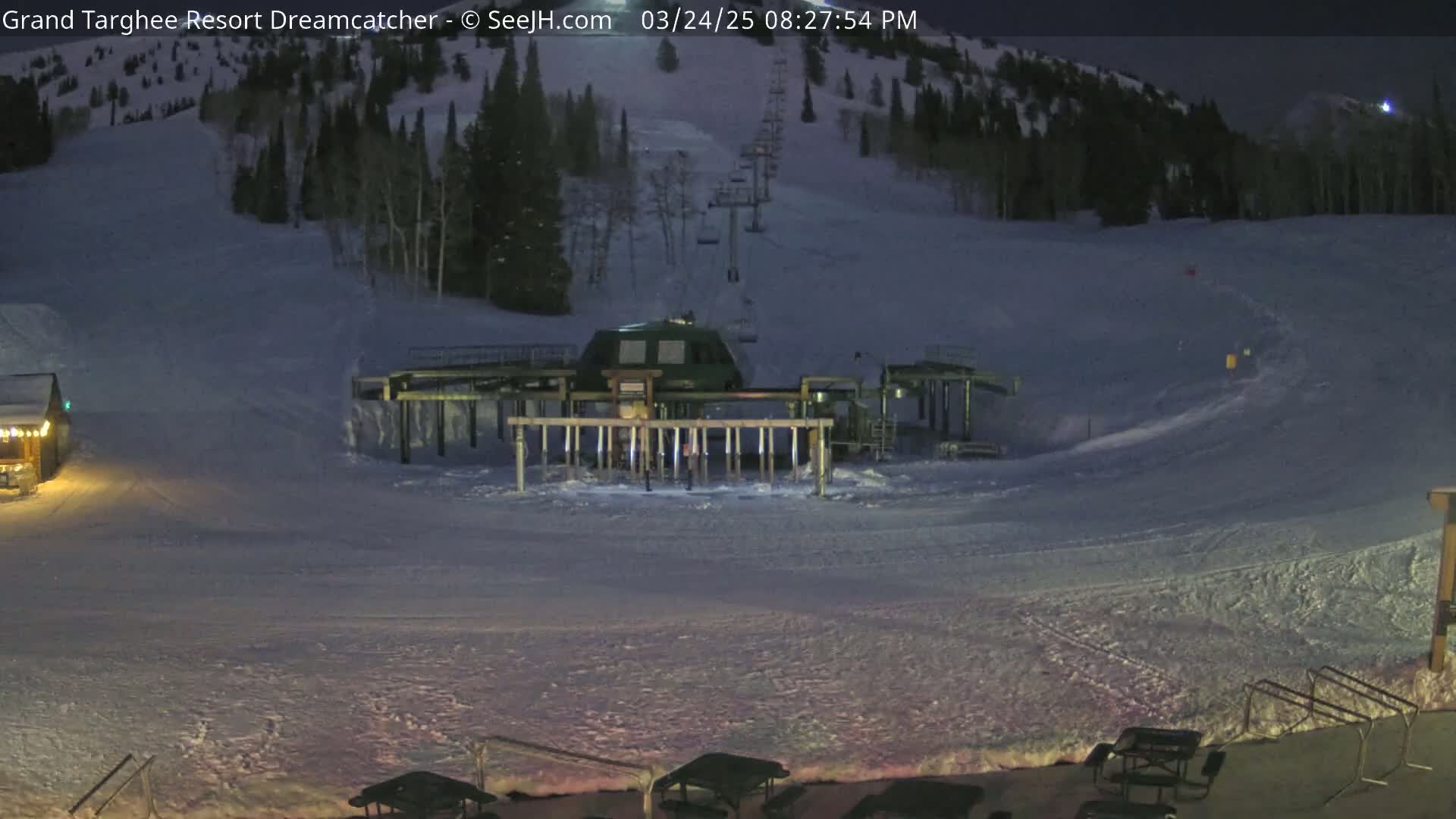 A ski lift base at night, under dark conditions, sits on a snow-covered slope with a ski run visible in the background.