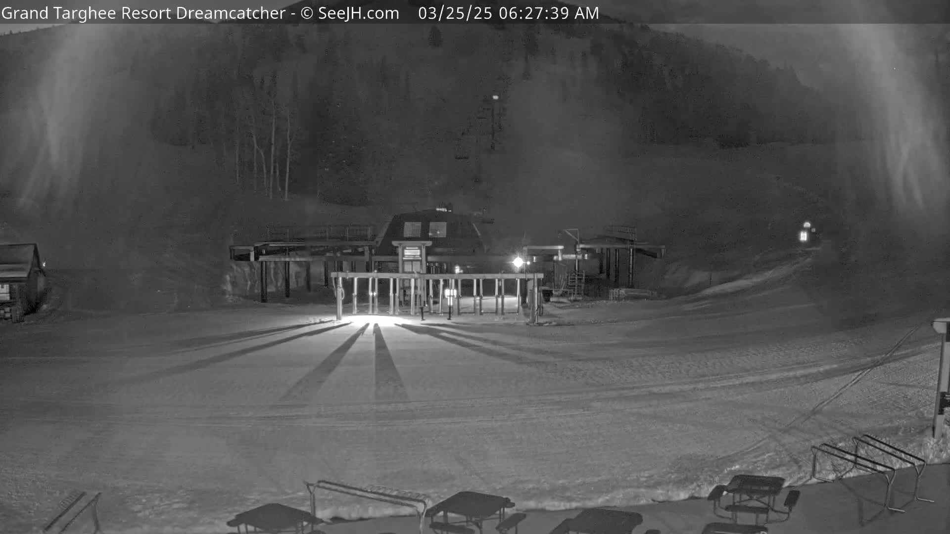 A snow-covered ski resort area at night, illuminated by lights, with a building and lift gates in the foreground and a snow-dusted hillside in the background under light snowfall.