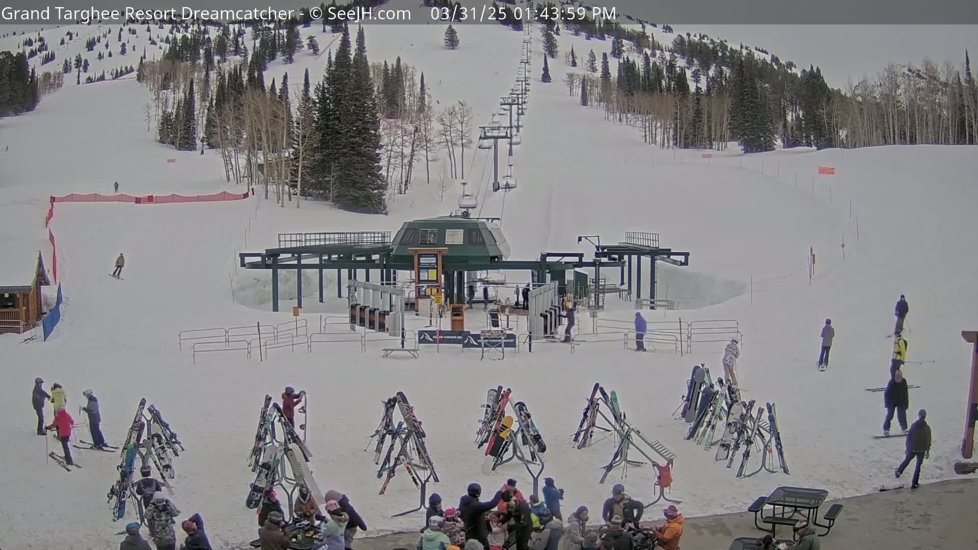 A ski lift base area with numerous pairs of skis on racks, surrounded by people, sits at the bottom of a snow-covered ski slope on a partly cloudy day.