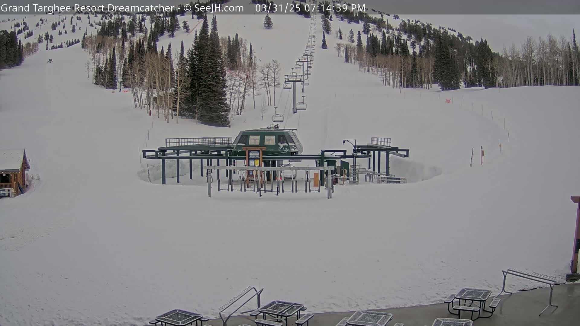 A ski lift base station sits in a snowy landscape, with a chairlift ascending a snow-covered hill lined with trees under an overcast sky.