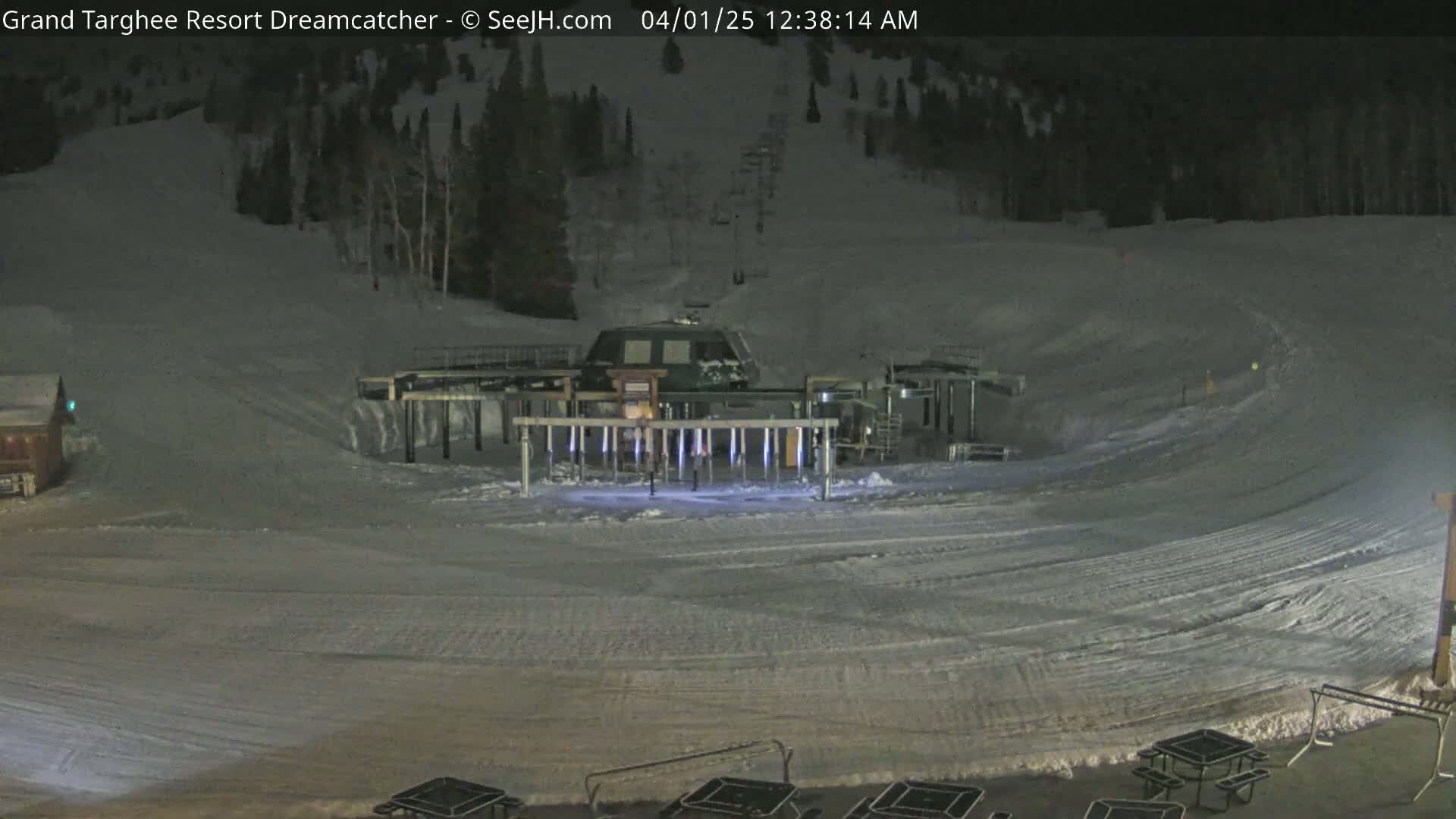 A ski lift base area at night, covered in snow, with a dark sky and a few picnic tables visible in the foreground.