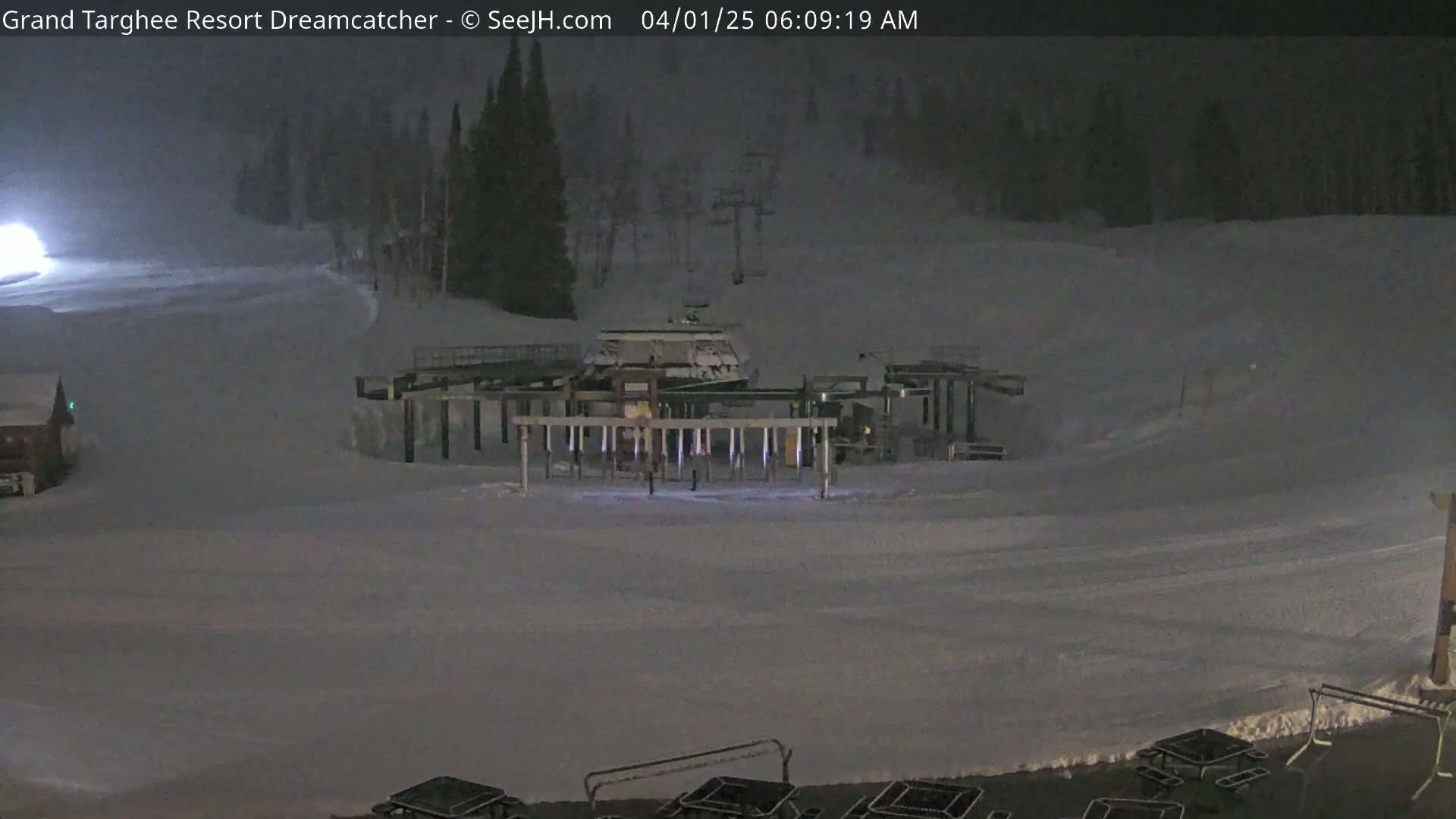 A nighttime, snow-covered ski resort scene shows a chairlift base and surrounding area, with a few structures and some picnic tables visible.