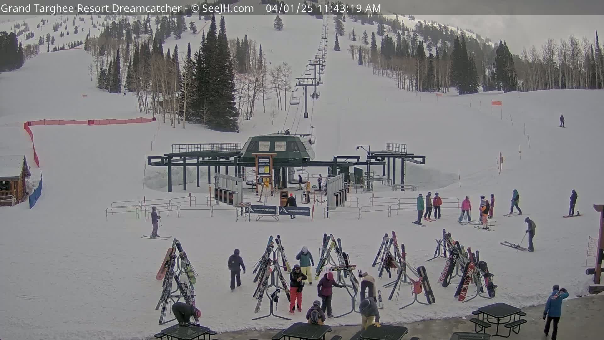 A ski resort base area with a chairlift, skiers and snowboarders, and several ski racks under an overcast sky.