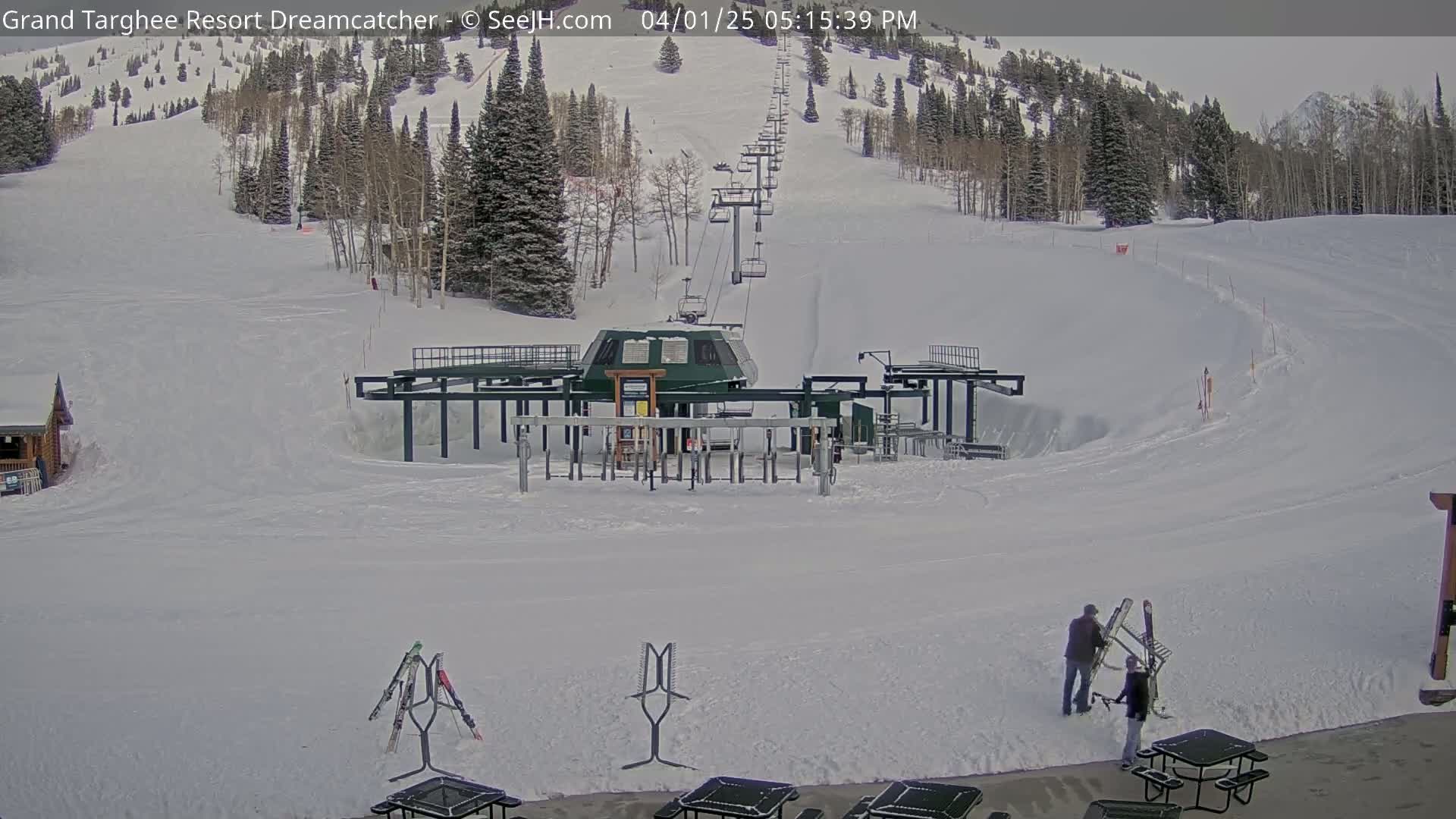 A ski lift base station sits at the bottom of a snow-covered ski slope, with two people near some picnic tables.