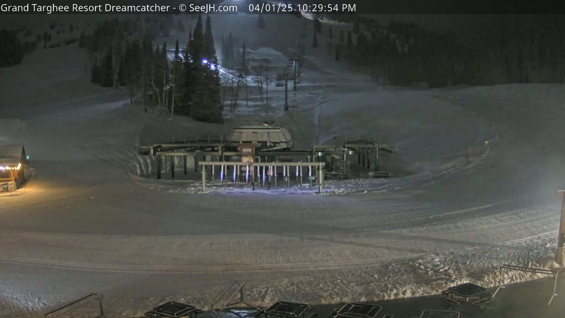 A nighttime view of a snow-covered ski resort with a chairlift and a base area showing snow-covered ground and some picnic tables.