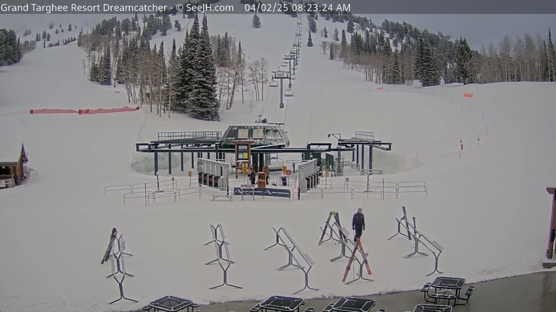 A snow-covered ski resort base area with a chairlift ascending a mountain, several ski racks, and a person walking near the lift terminal.