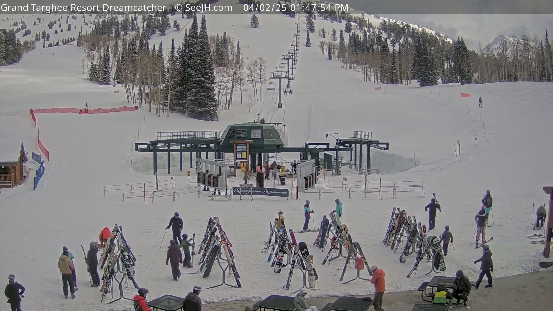 A ski lift base area with numerous skiers and snowboarders near racks of equipment, on a snowy day with overcast skies.