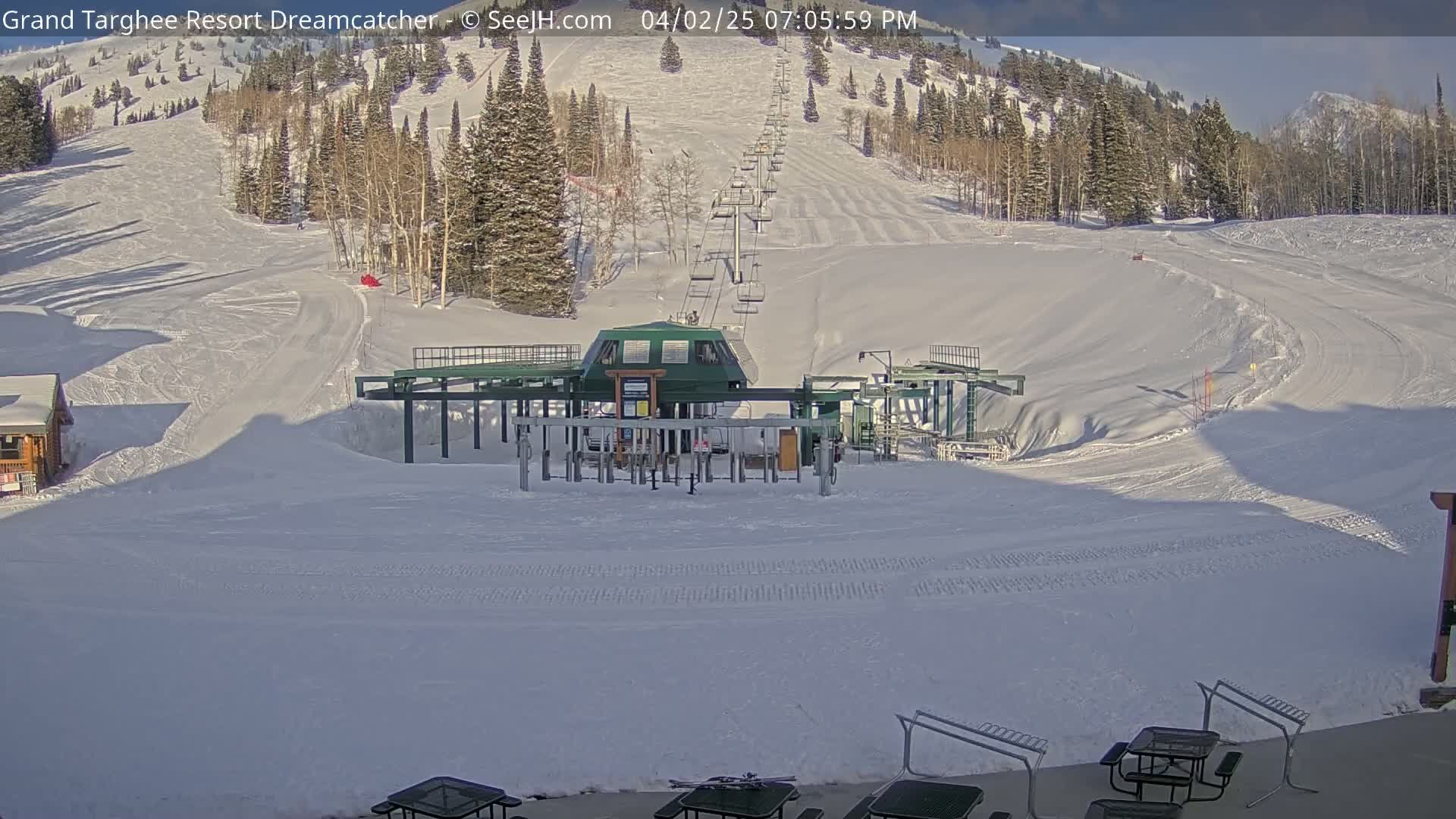 A ski lift base station sits at the bottom of a snow-covered ski slope on a sunny day, with mountains and trees in the background.