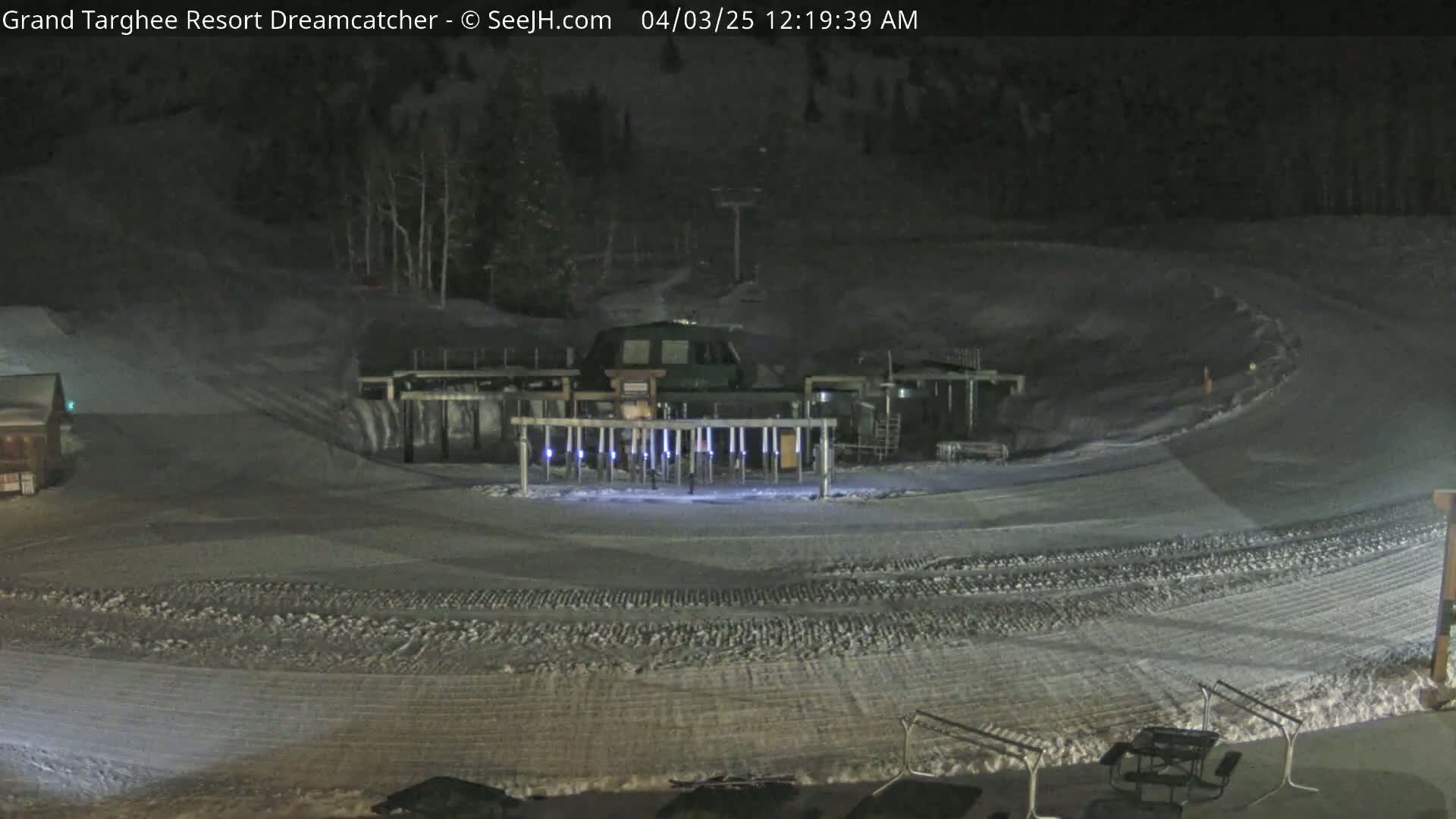 A nighttime view of a snow-covered ski slope with a lift mechanism and some outdoor furniture.