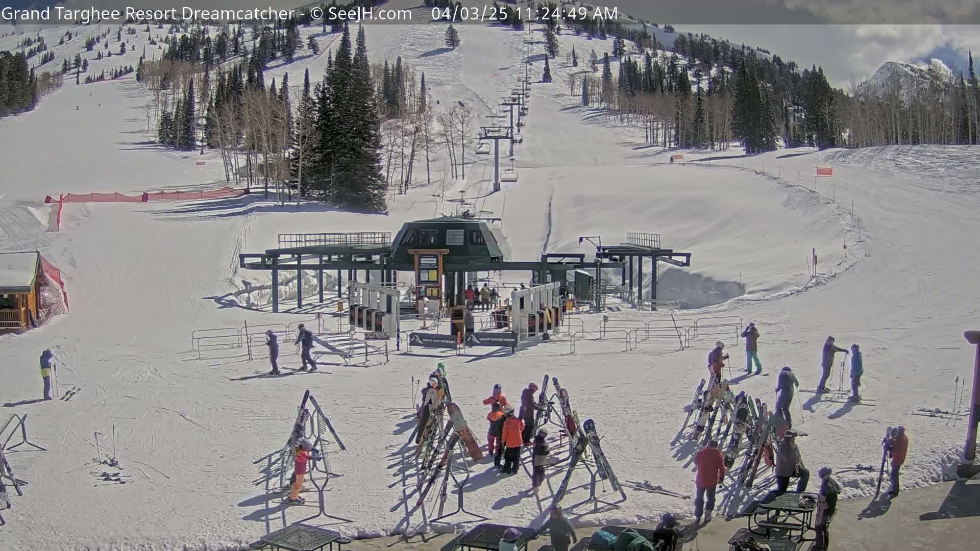 A sunny ski resort scene shows a ski lift base with skiers and snowboarders gathered around equipment racks, with snow-covered slopes and a mountain in the background.