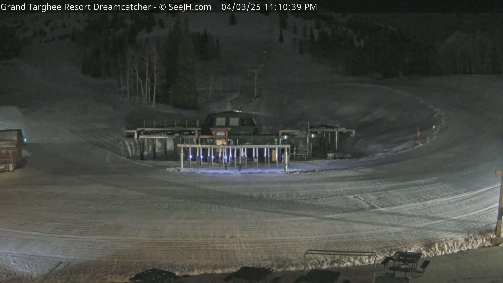 A snow-covered ski area at night, illuminated by lights around a ski lift base.