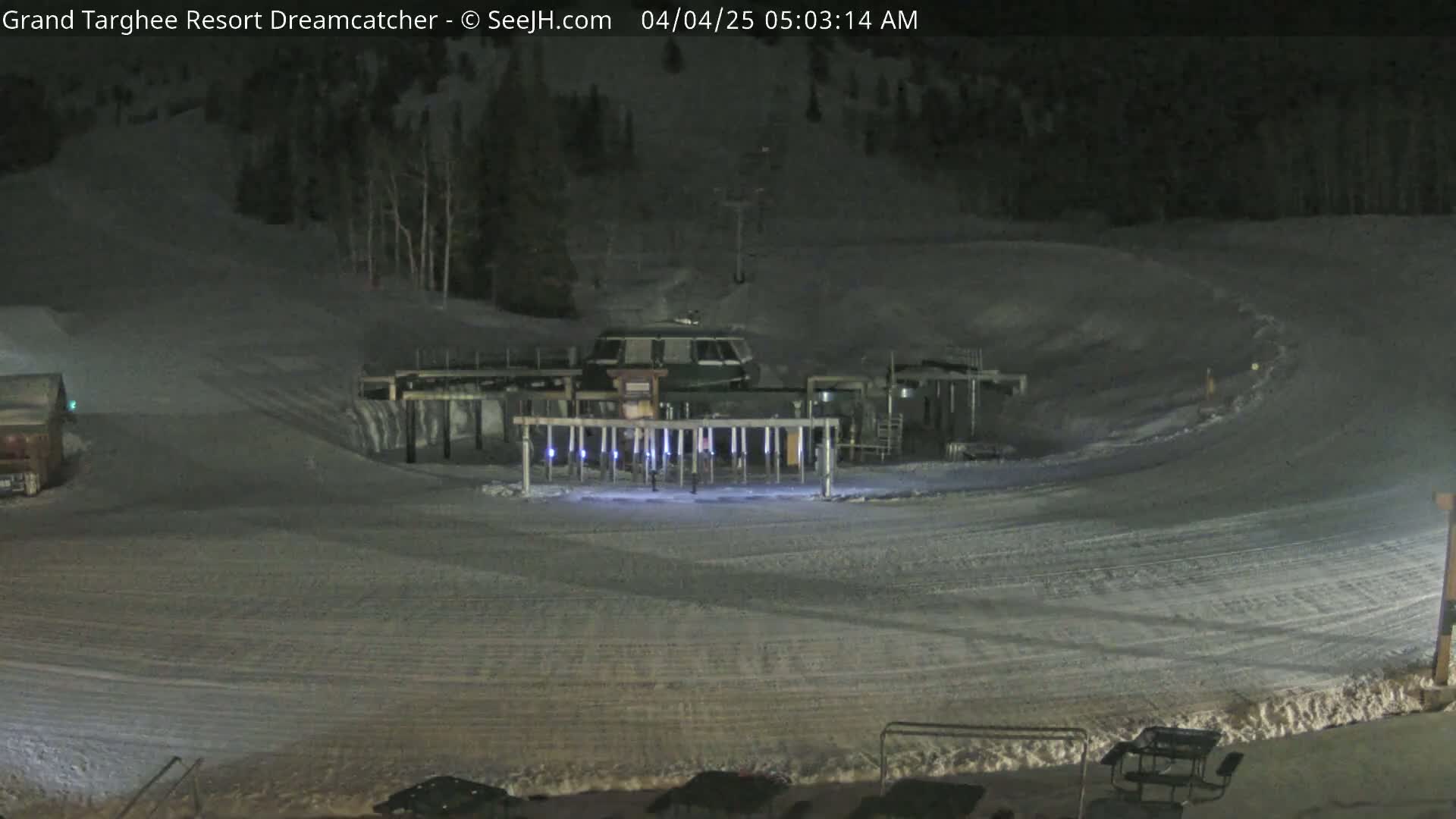 A snow-covered ski slope at night, illuminated by lights near a ski lift, with a dark forest in the background.