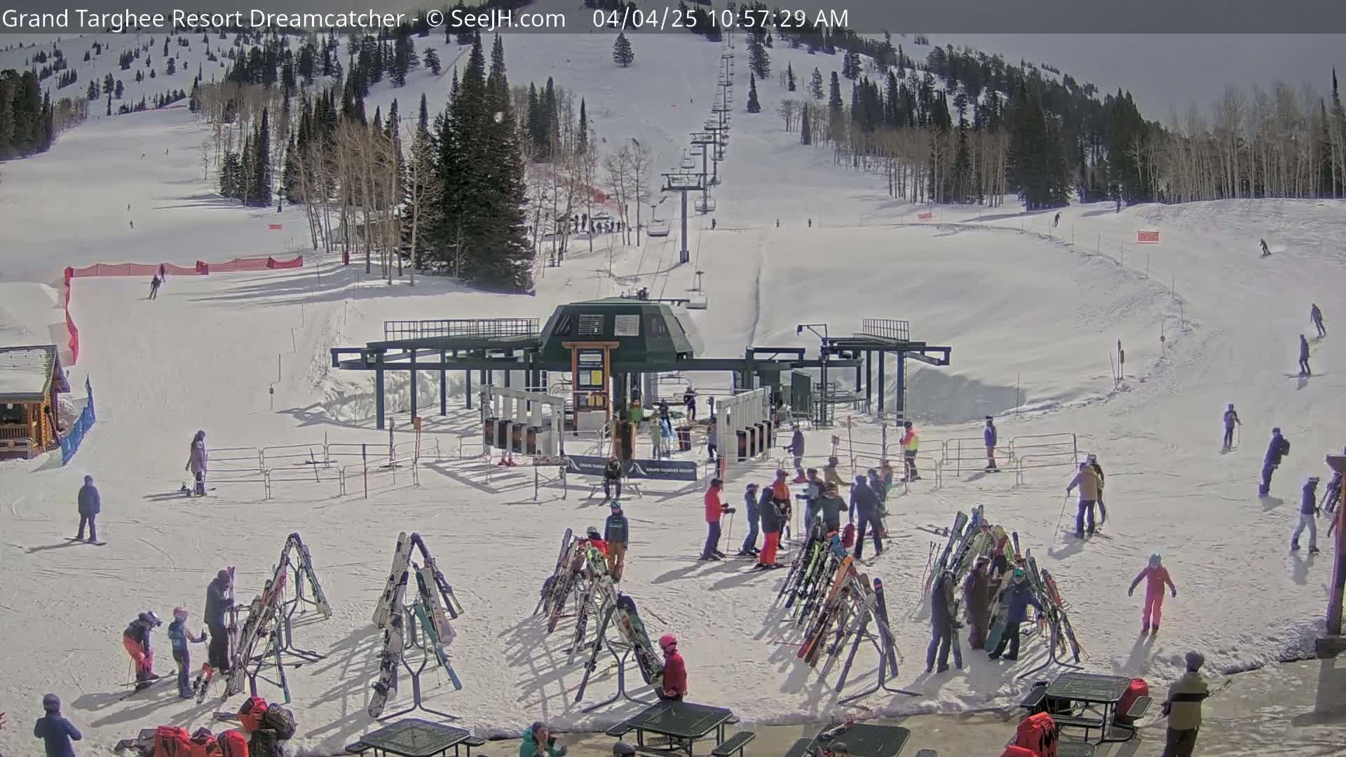 A sunny ski resort scene shows a ski lift, skiers and snowboarders on the slopes, and people gathering near ski racks at the base of a mountain.