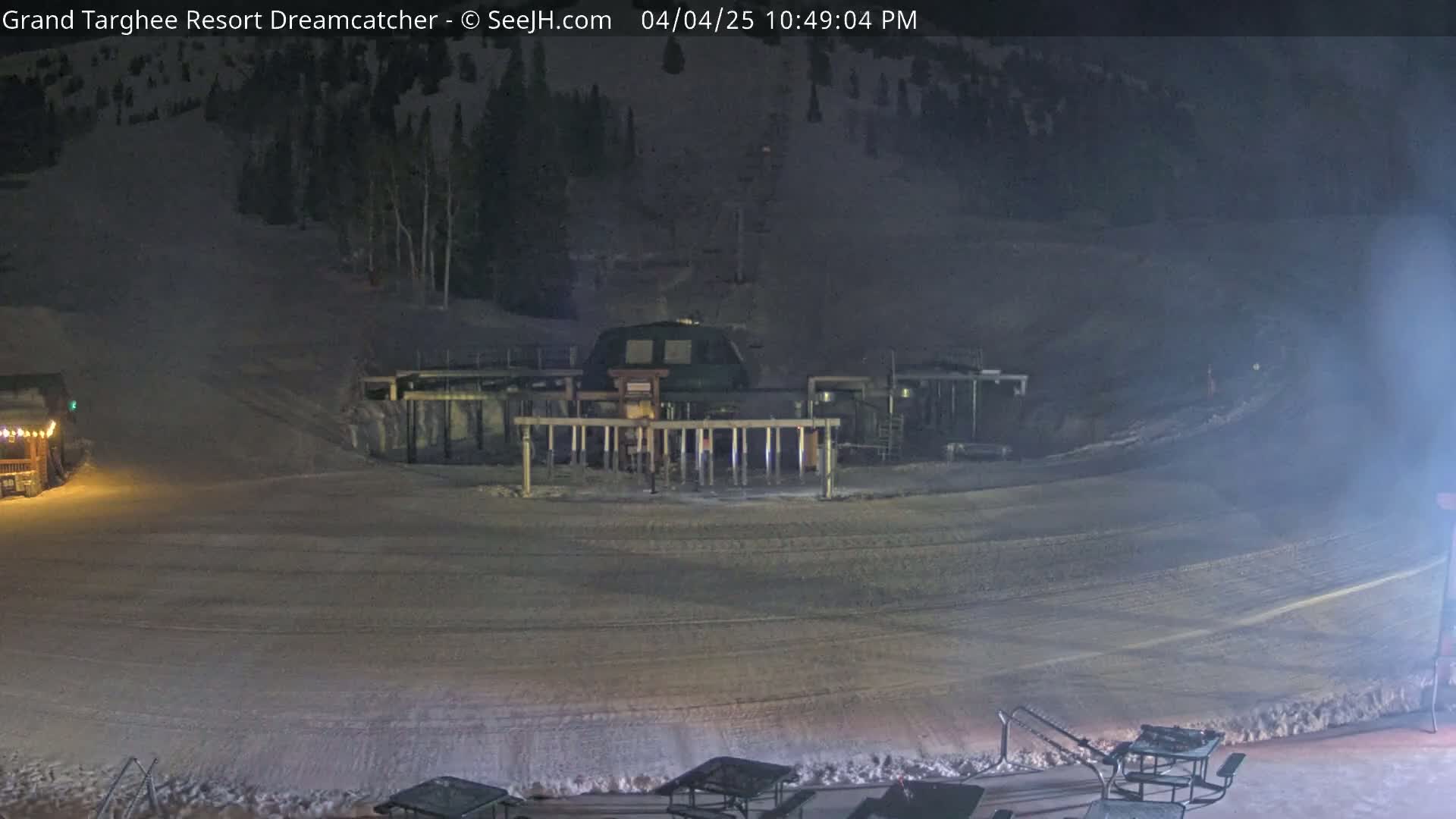 A nighttime view of a ski lift base area with snow-covered ground and a few outdoor tables.