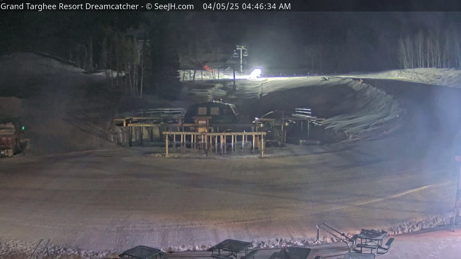 A nighttime view of a snow-covered ski slope with a snow groomer and ski lift visible, under dark conditions.