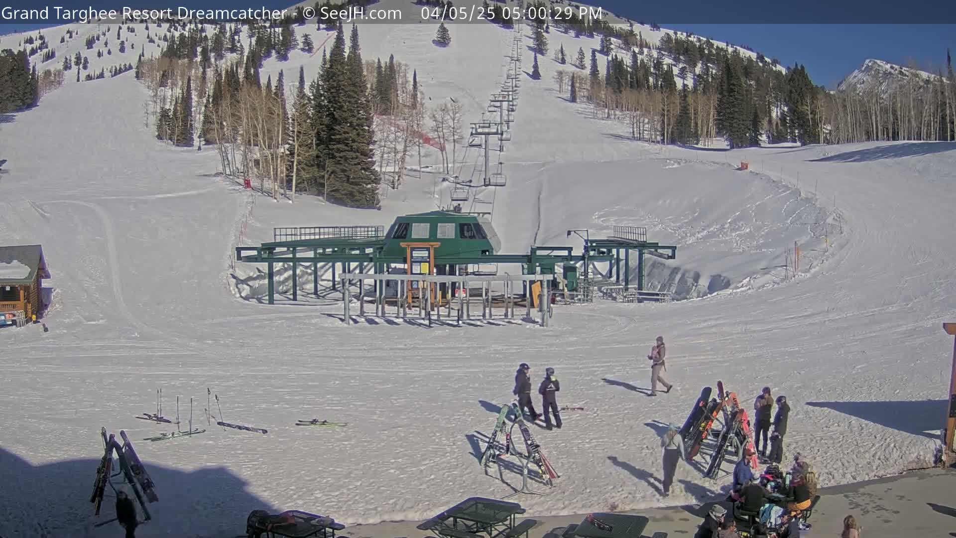 A ski lift base area with people standing around, skis leaning against a building, and a sunny, snow-covered mountain slope in the background.