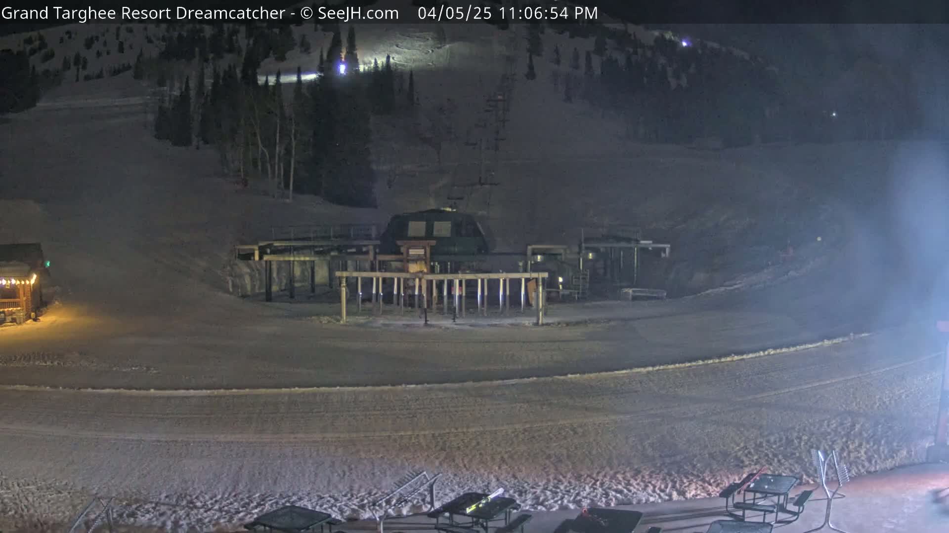 A snow-covered ski resort at night, showing a ski lift and a few outdoor tables.
