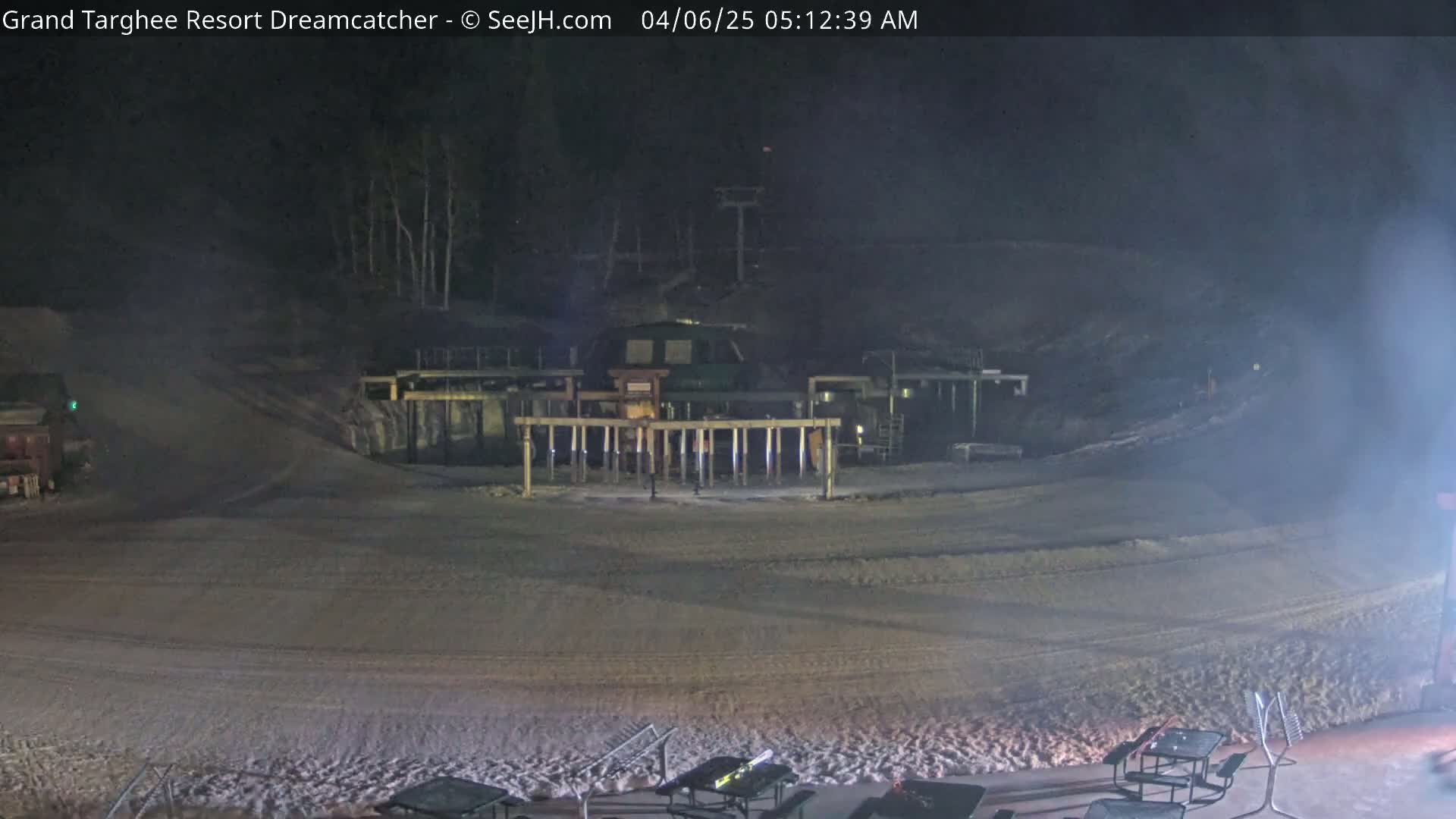A nighttime view of a snow-covered ski resort area shows a ski lift and a building with lights, under hazy conditions.