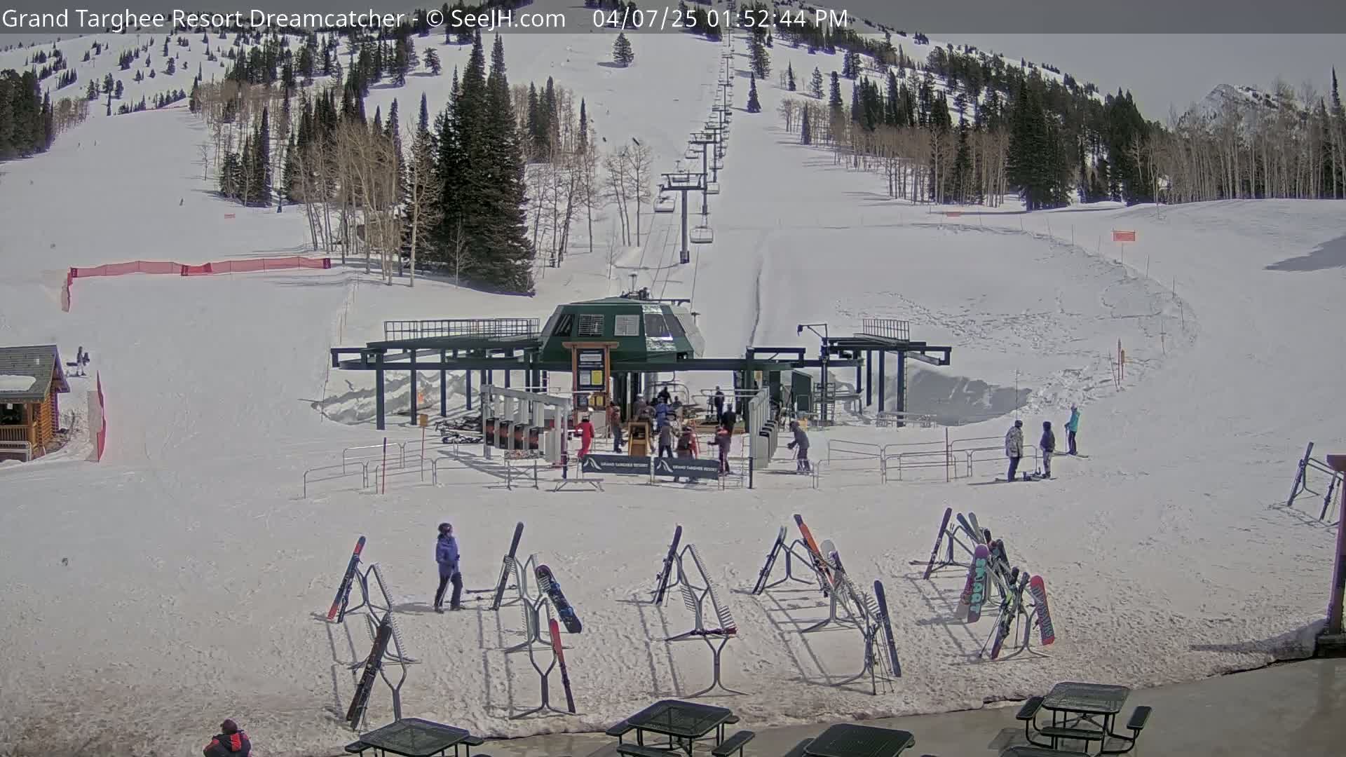 A ski lift base area with several people and skis on racks, under a sunny sky, on a snowy mountainside.