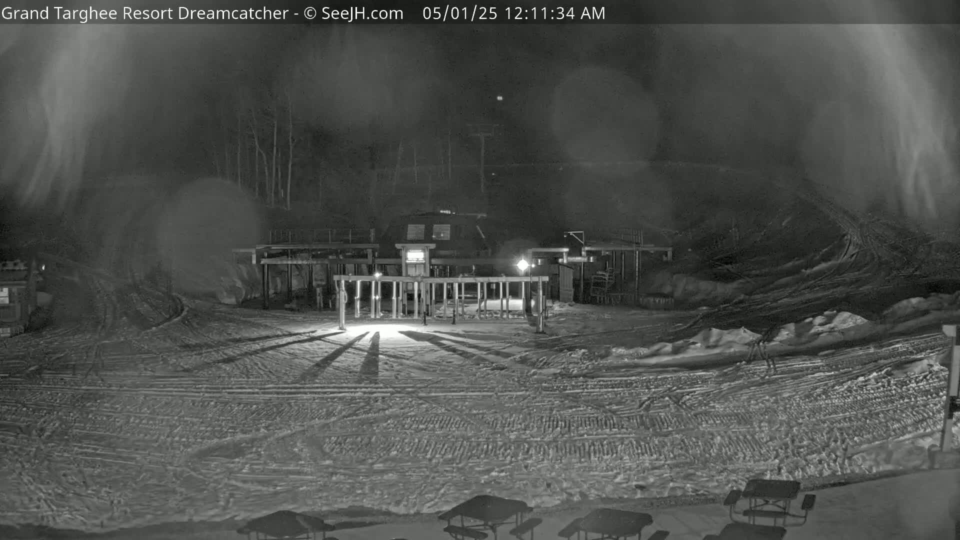 A snow-covered nighttime scene shows a building with lights and a gated area, with shadows cast on the snow-covered ground under a hazy sky.