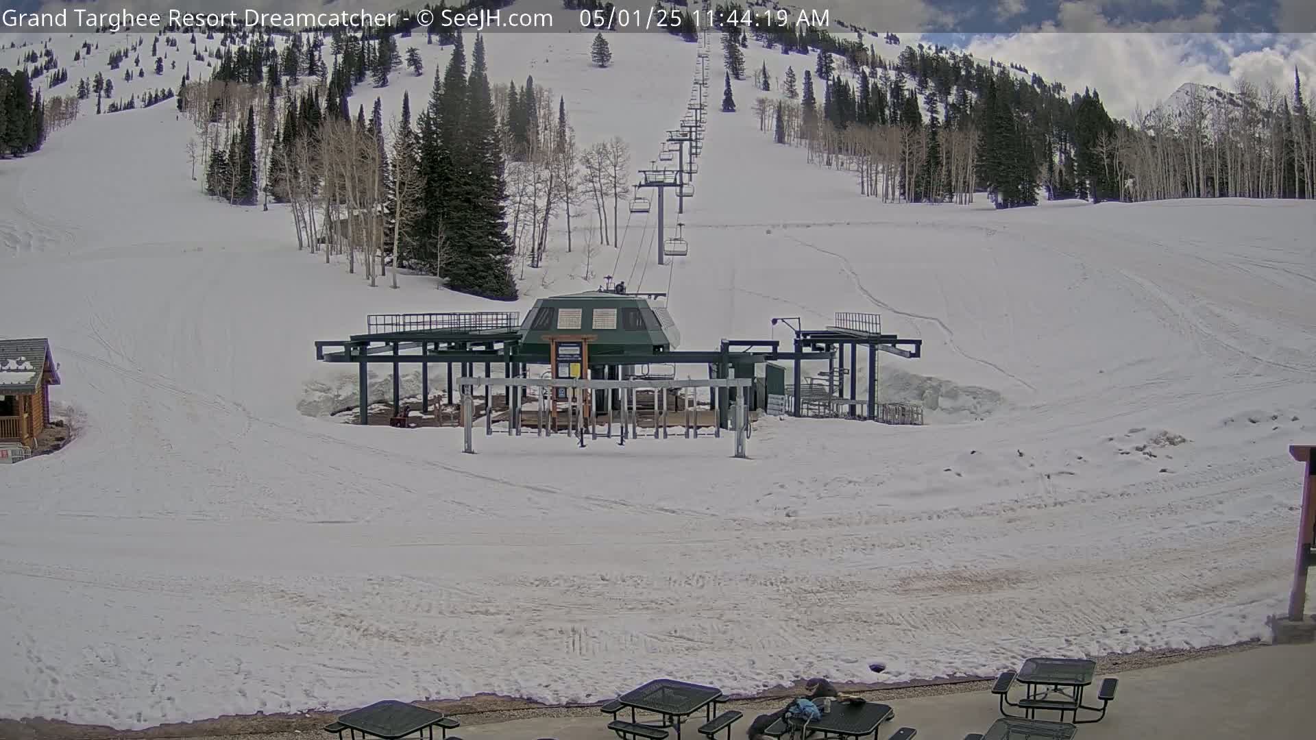 A ski lift base station sits on a snow-covered slope under a partly cloudy sky, with mountains in the background.