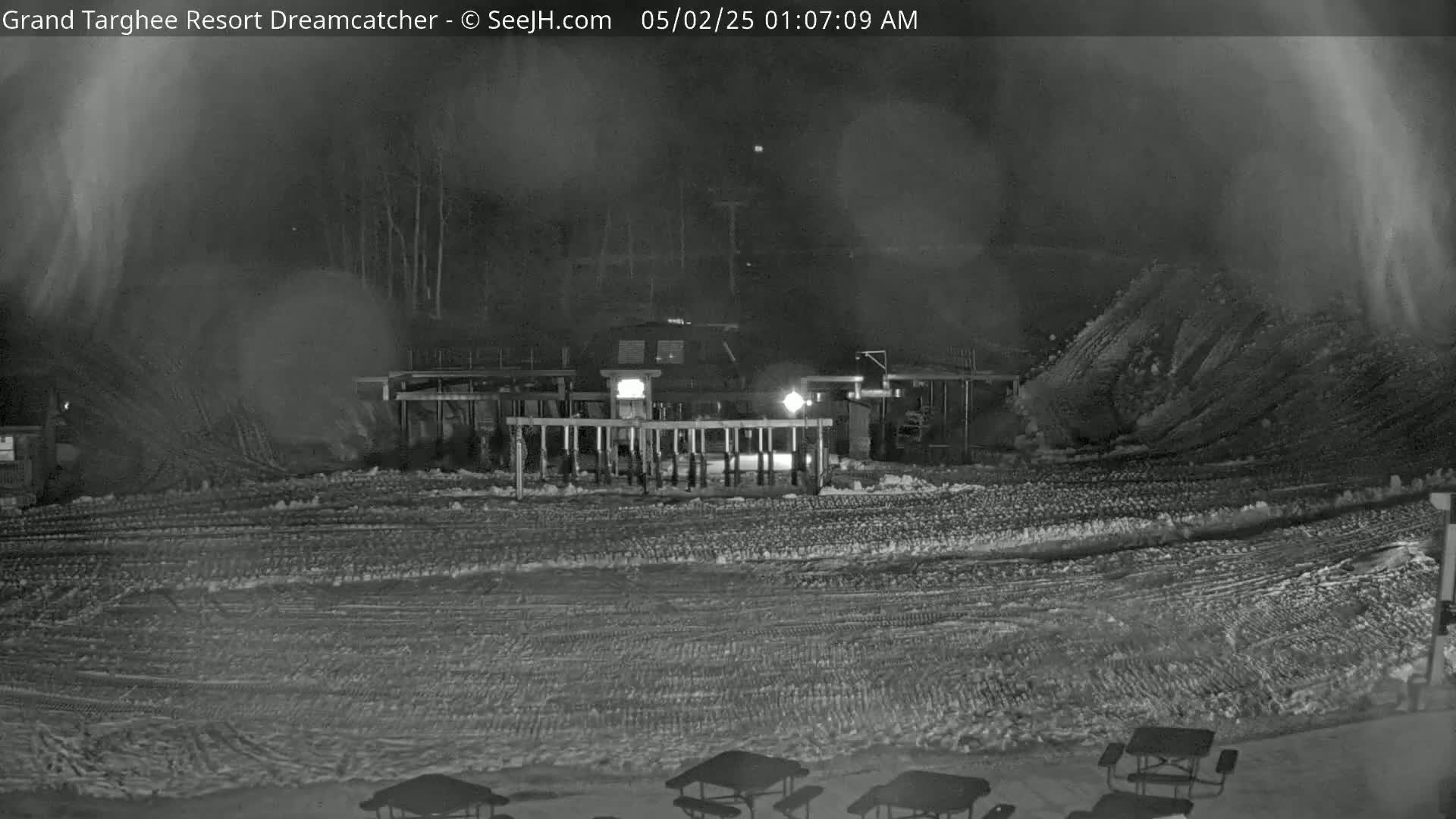 A nighttime, snow-covered scene shows a building with a fence and several picnic tables, under hazy conditions.