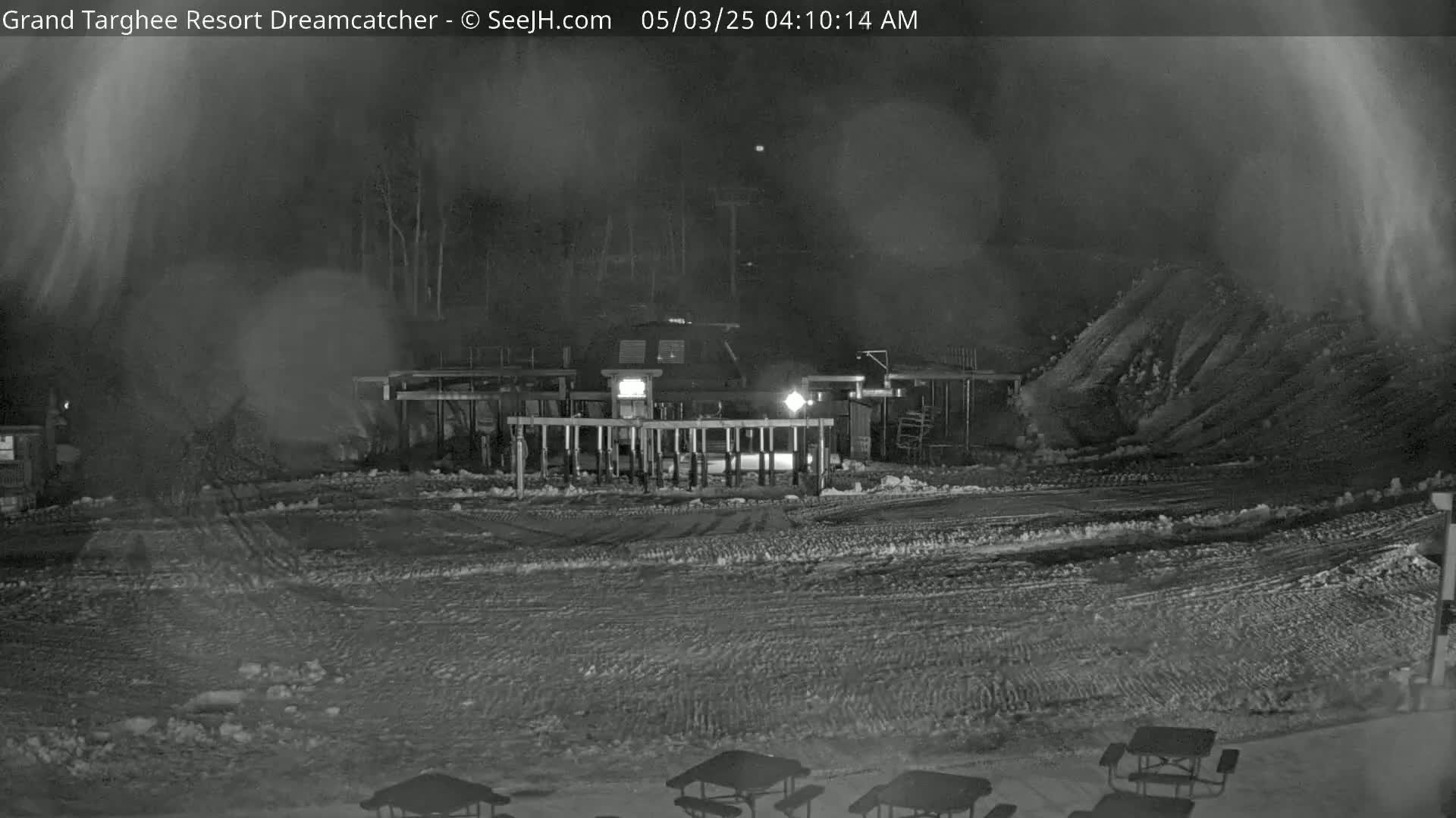 A nighttime snow scene shows a snow-covered area with a building and several picnic tables, with blowing snow in the foreground.