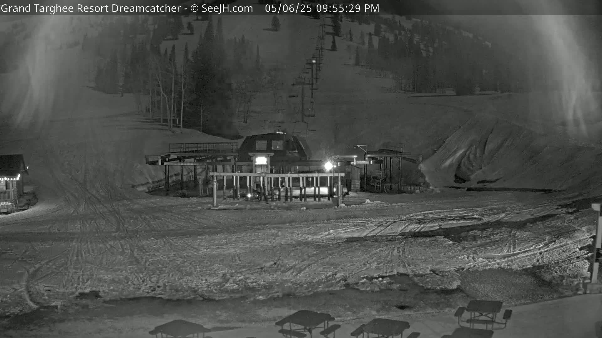 A nighttime view of a ski lift base area with snow-covered ground and a few picnic tables.