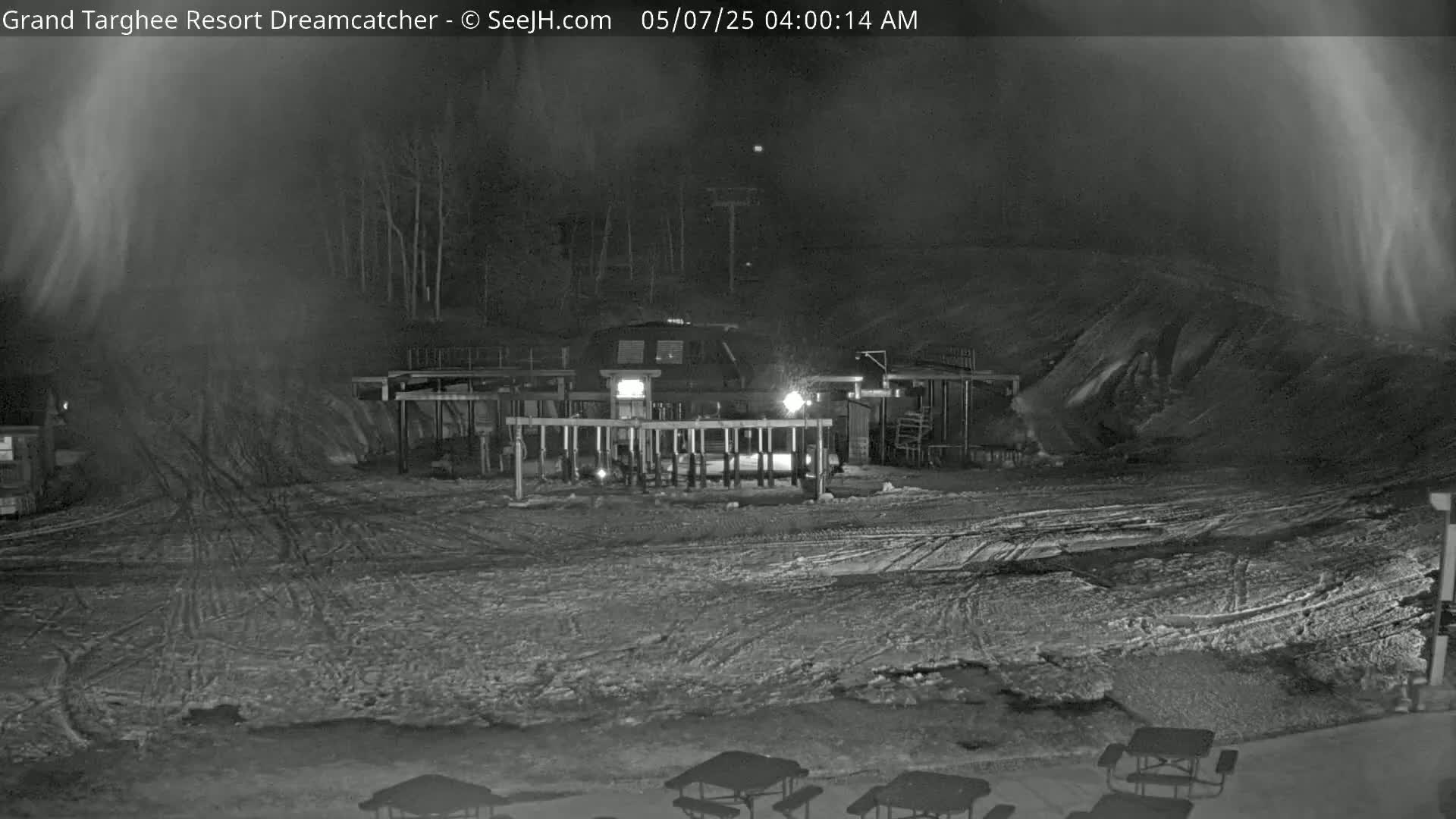 A nighttime snow-covered scene shows a ski lift building with a gate and several picnic tables in the foreground, under hazy conditions.