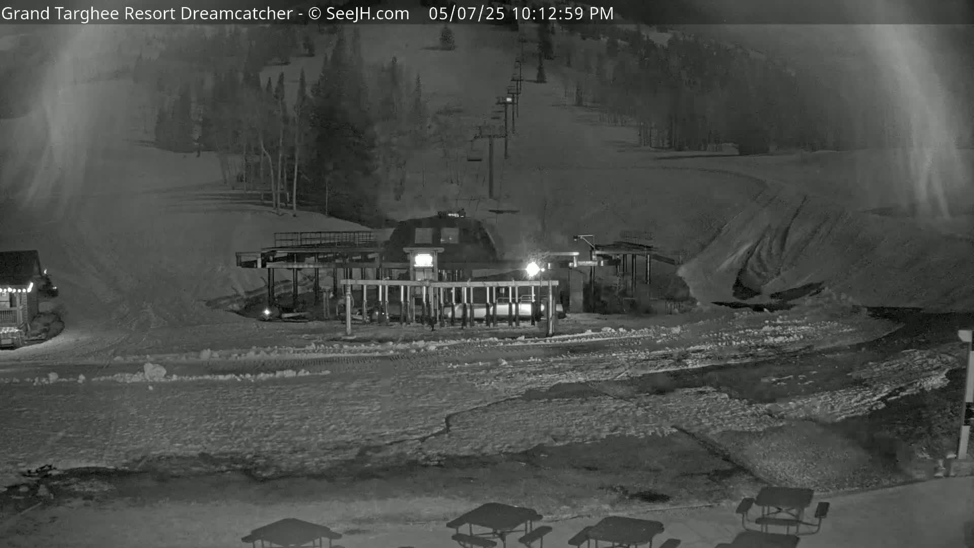 A nighttime, snow-covered ski resort scene shows a ski lift base station with a chairlift extending uphill, under a partly cloudy sky.