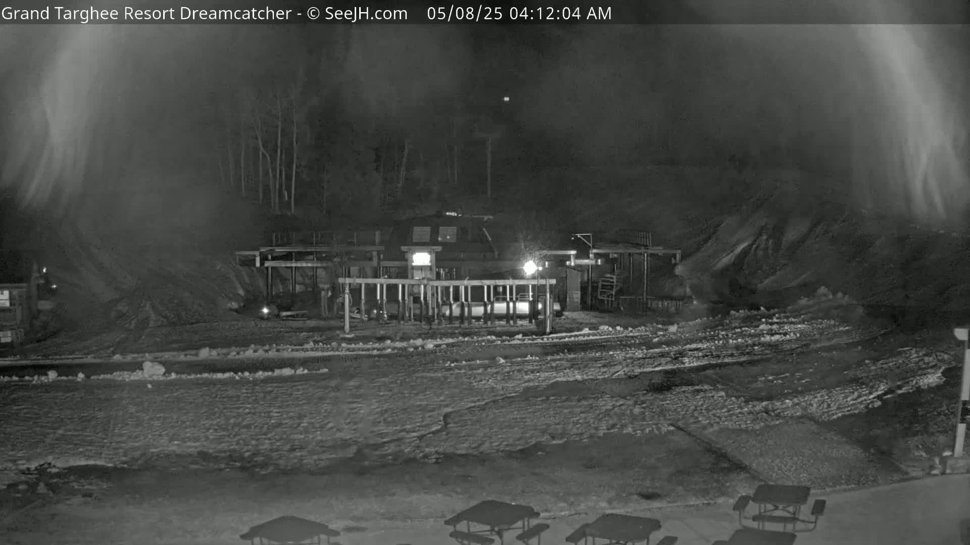 A nighttime, snow-covered scene shows a building with lights on, surrounded by a snowy area with picnic tables.