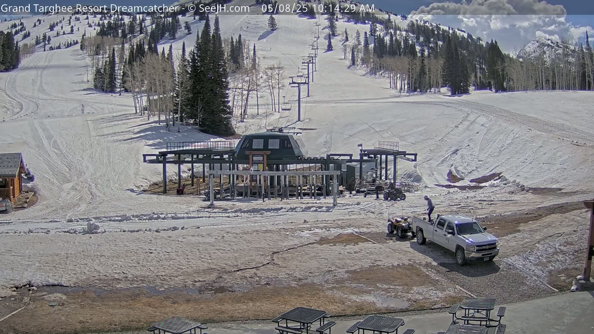 A ski lift base station sits in a snowy mountain area under a partly sunny sky, with a pickup truck and several all-terrain vehicles parked nearby.
