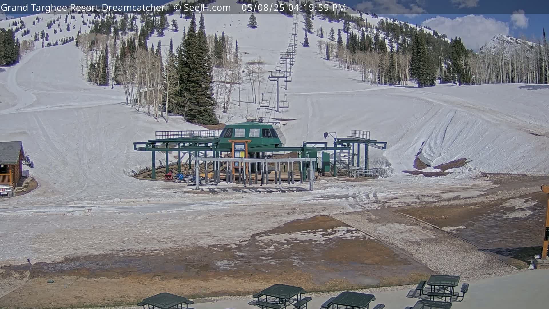 A ski lift base station sits on melting snow at the bottom of a snow-covered mountain slope under a partly sunny sky.