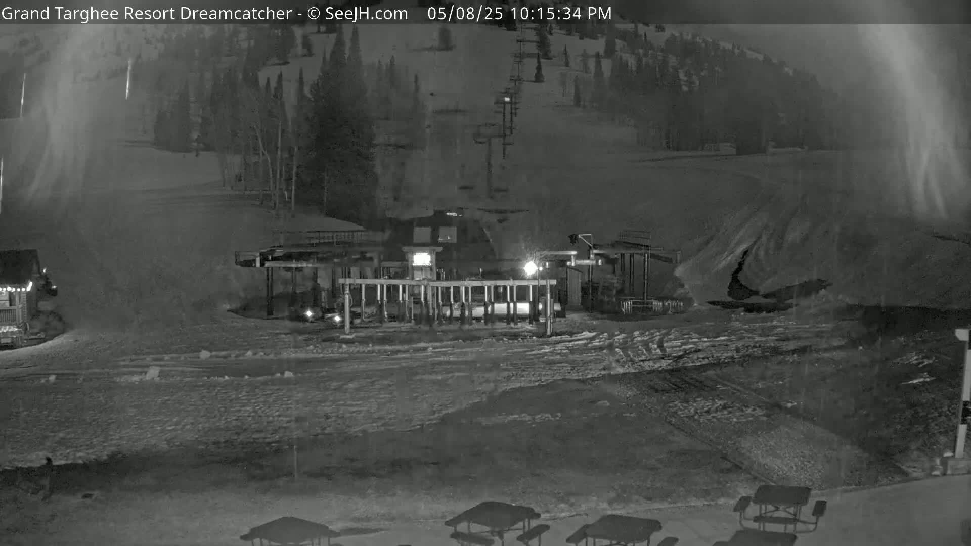 A nighttime, snowy scene shows a ski lift base station and snow-covered terrain, with a few picnic tables visible in the foreground.