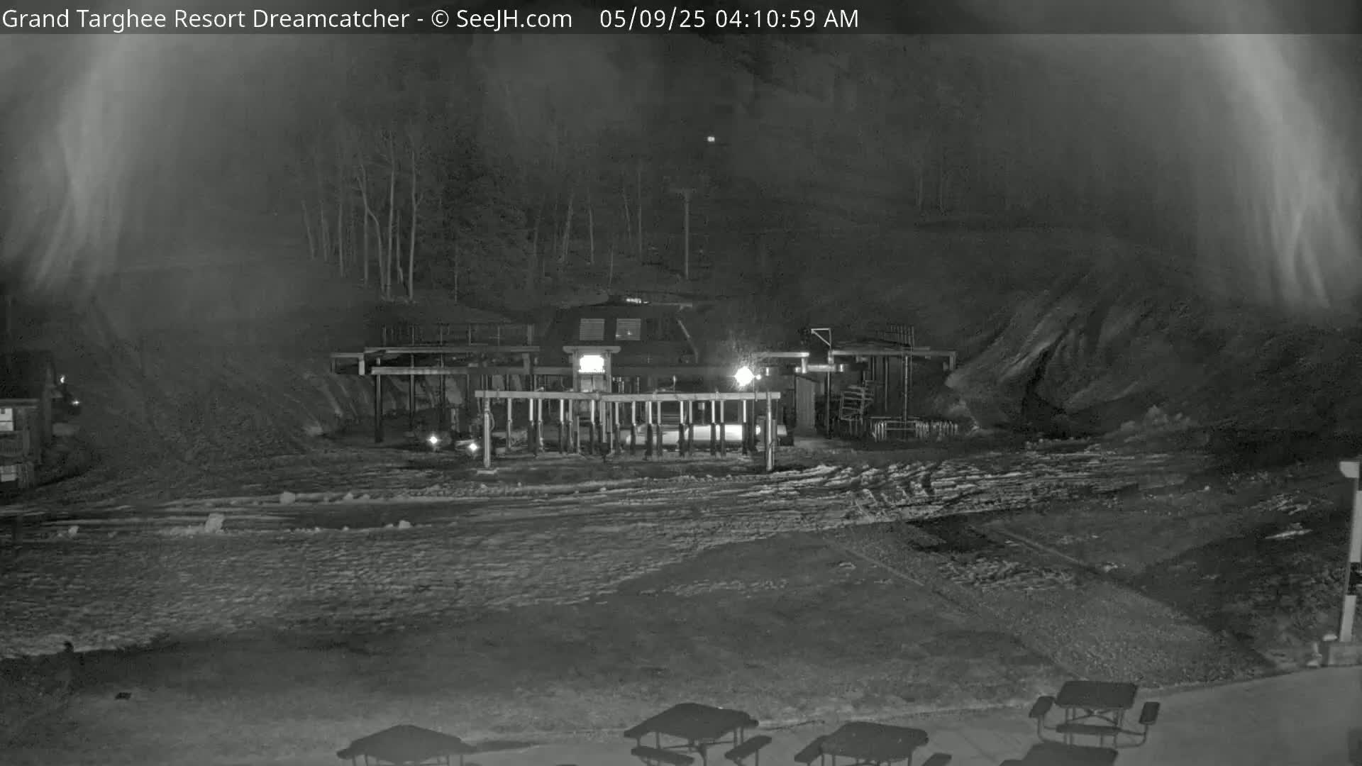 A nighttime, snow-covered scene shows a ski lift station with a building and several picnic tables in the foreground.