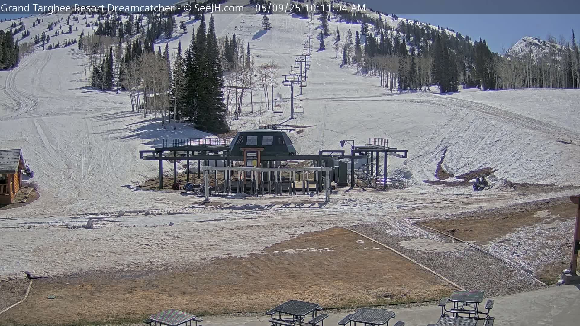 A ski lift base station sits at the bottom of a snow-covered ski slope on a sunny day, with mountains visible in the background.