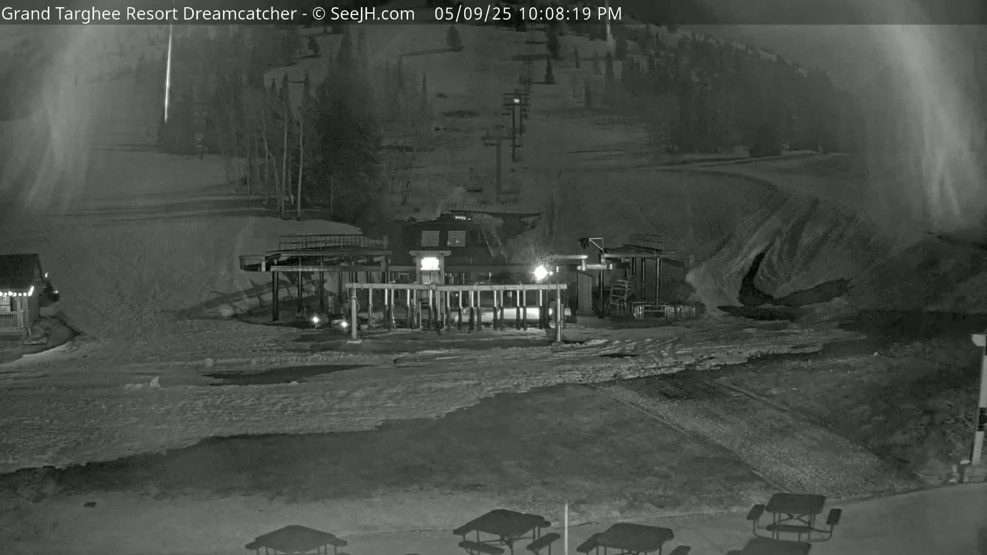 A nighttime, snow-covered ski resort scene shows a lift station with lights on, picnic tables, and a building in the background under a dark, possibly cloudy sky.
