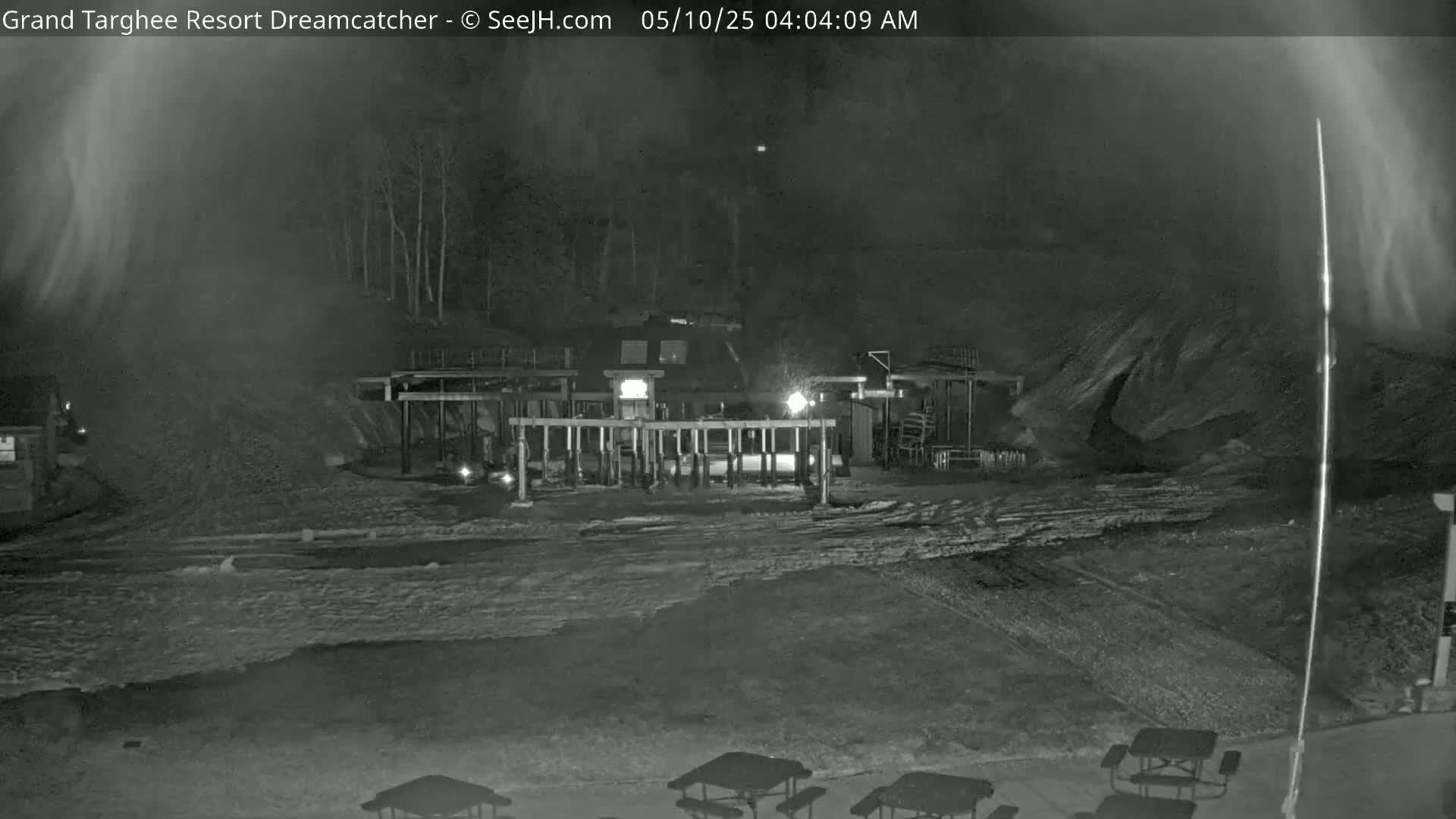 A nighttime, snowy scene shows a building with lights on and picnic tables in a snow-covered area.