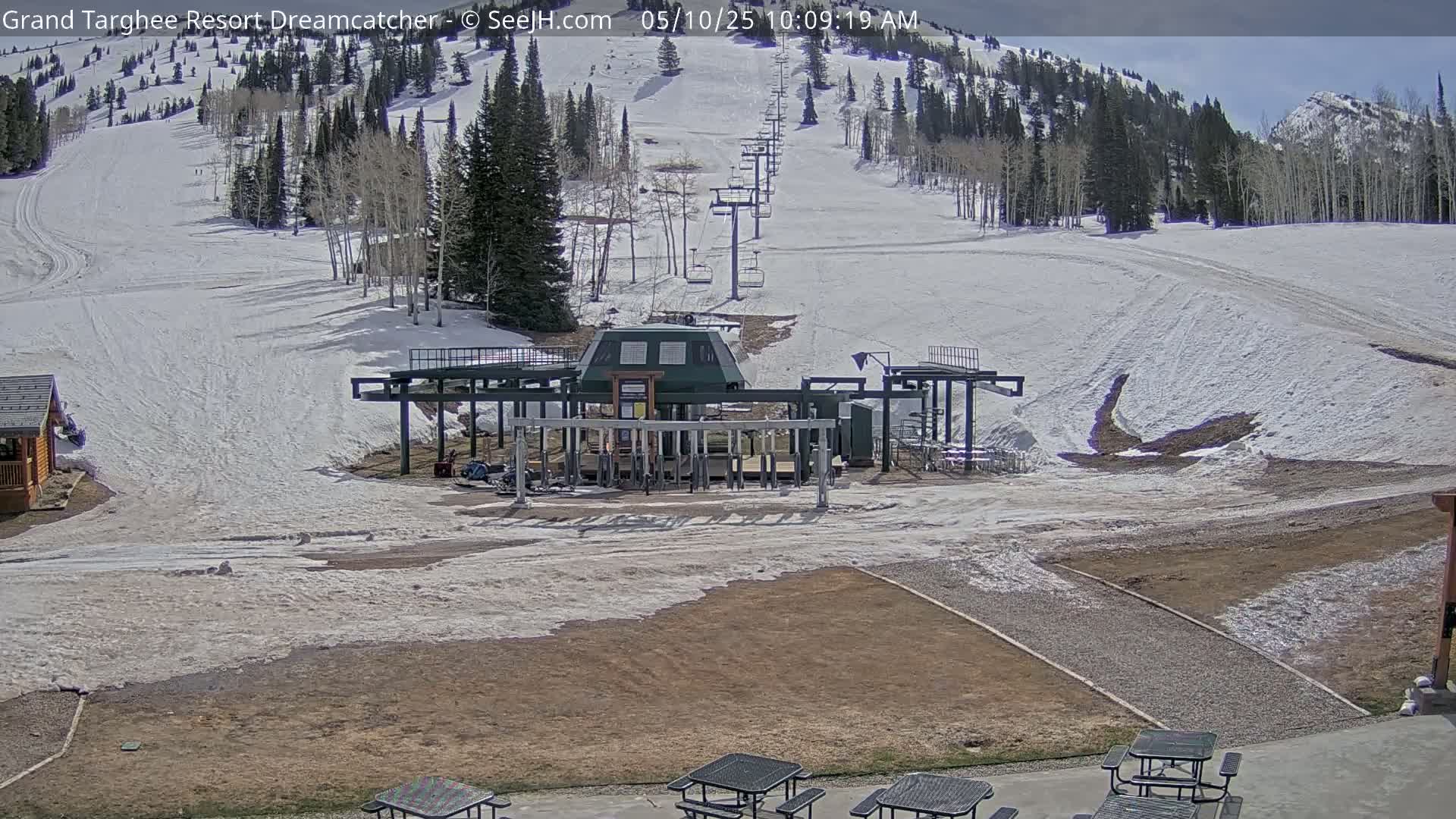 A ski lift base station sits at the bottom of a snow-covered ski slope on a sunny day, with mountains in the background and picnic tables in the foreground.