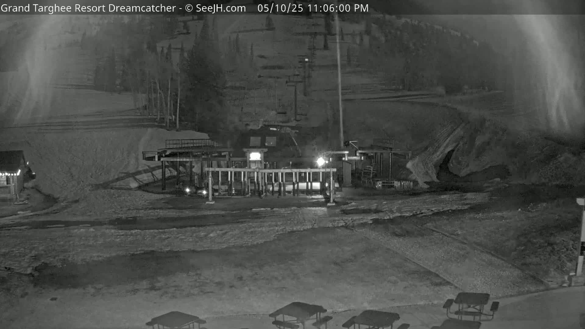 A nighttime, snow-covered ski resort scene shows a lift station with lights on, a small building, and several picnic tables in the foreground.