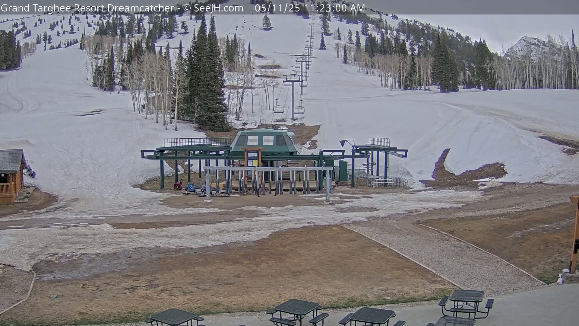 A ski lift base station sits at the bottom of a snow-covered ski slope, with patchy snow and bare ground visible in the foreground, under an overcast sky.