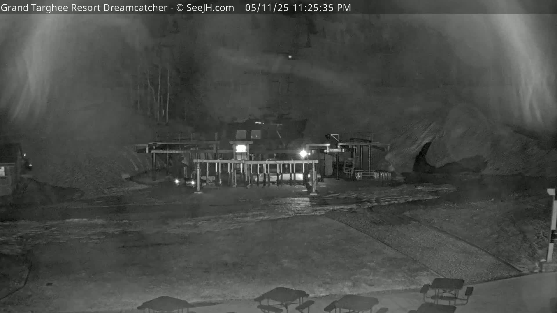 A nighttime, low-light view shows a snow-covered area with a structure and several picnic tables, under a mostly cloudy sky.